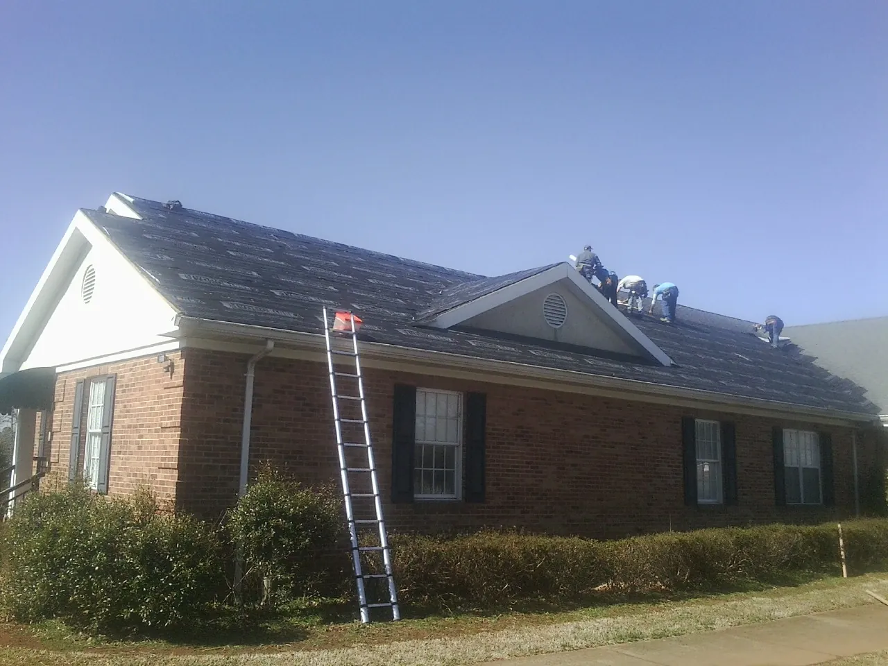 Crew tearing shingles off a brick ranch roof with a ladder set — Emergency Roof Repair work by our crew in Cement