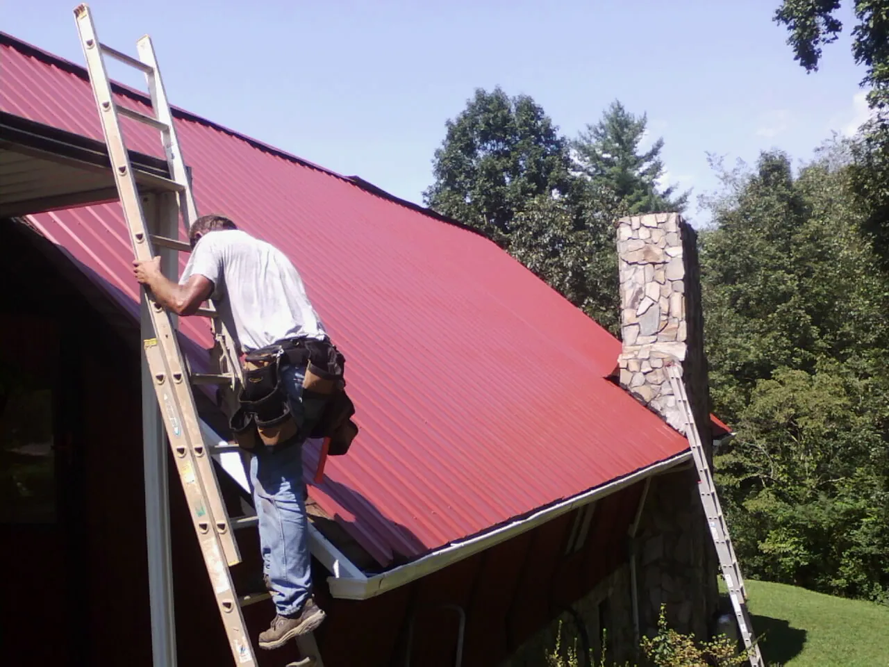 Roofer on a ladder beside a red metal roof and stone chimney — Soffit & Fascia Repair by Roofing Brothers in Cement